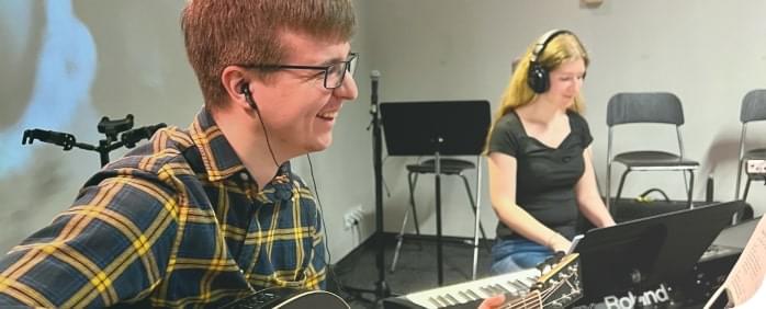 uśmiechnięty chłopak grający na gitarze, a w tle dziewczyna ze słuchawkami grająca na keyboardzie